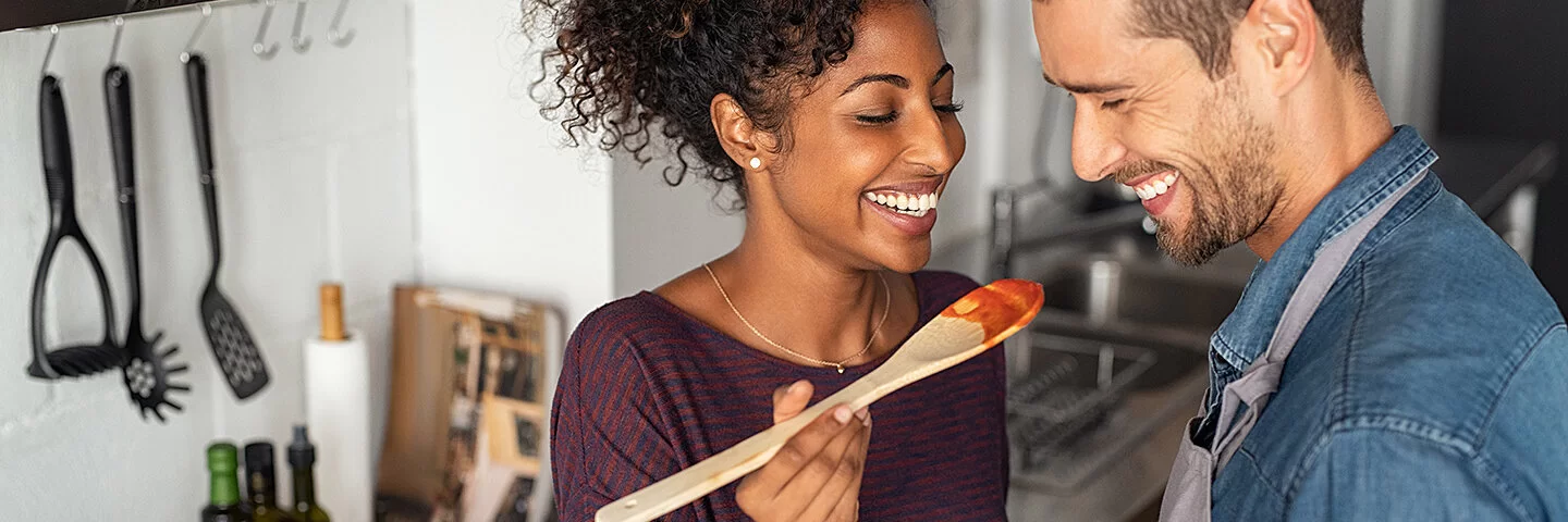 Gesund und günstig kochen: Eine Frau und ein Mann stehen gemeinsam in der Küche und schauen lachend auf einen Kochlöffel mit Tomatensoße.