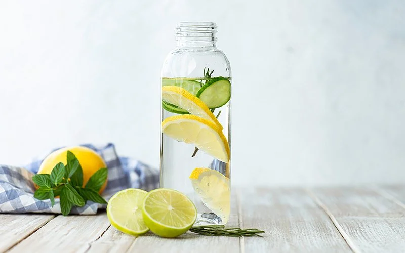 Flasche mit Infused Wasser auf weißem Holz mit einer Scheibe Zitrone, Gurke und Rosmarinblatt darin.
