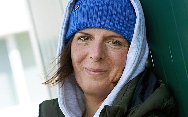 Die Busfahrerin Angela Rossberg lehnt draußen lässig an einer dunkelgrünen Wand. Sie trägt eine blaue Mütze und darüber die Kapuze ihres grauen Hoodies, sie lächelt in die Kamera.