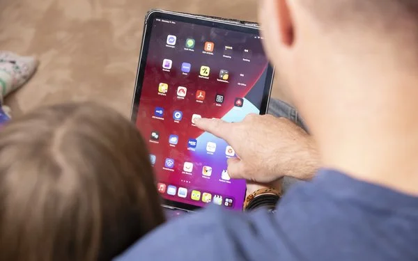 Vater und Tochter sind von hinten zu sehen, wie sie gemeinsam an einem Tablet sitzen und eine App auswählen.