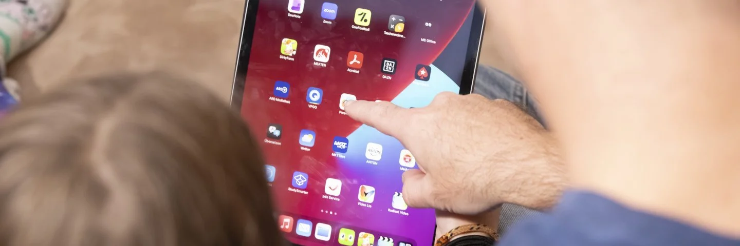 Vater und Tochter sind von hinten zu sehen, wie sie gemeinsam an einem Tablet sitzen und eine App auswählen.