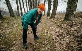 Ein älterer Mann hat beim Spaziergang auf einem Feldweg Atemnot und stützt sich mit den Händen auf den Knien ab.