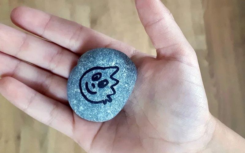 Eine offene Hand hält einen grauen Stein mit einer Zeichnung von einem lächelnden Drachenkopf.