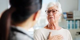 Eine ältere Person mit kurzen, weißen Haaren sitzt in einem hellen Raum und trägt einen beigen Pullover. Die Person hält eine Hand auf die Brust. Im Vordergrund ist eine weitere Person von hinten zu sehen, die einen weißen Kittel und ein Stethoskop um den Hals trägt. Im Hintergrund stehen ein Stuhl und medizinische Geräte.