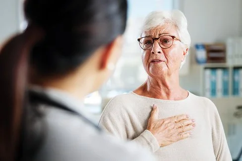 Eine ältere Person mit kurzen, weißen Haaren sitzt in einem hellen Raum und trägt einen beigen Pullover. Die Person hält eine Hand auf die Brust. Im Vordergrund ist eine weitere Person von hinten zu sehen, die einen weißen Kittel und ein Stethoskop um den Hals trägt. Im Hintergrund stehen ein Stuhl und medizinische Geräte.