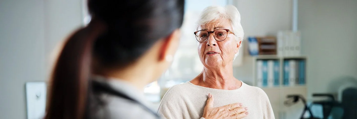 Eine ältere Person mit kurzen, weißen Haaren sitzt in einem hellen Raum und trägt einen beigen Pullover. Die Person hält eine Hand auf die Brust. Im Vordergrund ist eine weitere Person von hinten zu sehen, die einen weißen Kittel und ein Stethoskop um den Hals trägt. Im Hintergrund stehen ein Stuhl und medizinische Geräte.