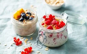 Auf einem Tisch stehen zwei geöffnete Gläser mit Overnight Oats. Als  Topping dienen Himbeeren, rote Johannisbeeren, Blaubeeren und ein Stück Mango.
