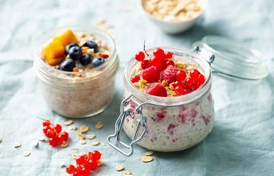 Auf einem Tisch stehen zwei geöffnete Gläser mit Overnight Oats. Als  Topping dienen Himbeeren, rote Johannisbeeren, Blaubeeren und ein Stück Mango.