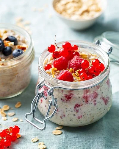 Auf einem Tisch stehen zwei geöffnete Gläser mit Overnight Oats. Als  Topping dienen Himbeeren, rote Johannisbeeren, Blaubeeren und ein Stück Mango.