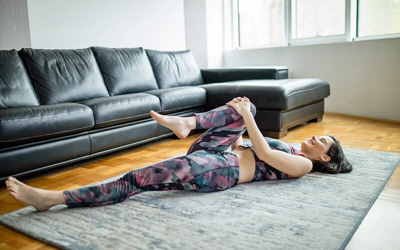 Junge Frau liegt in Sportkleidung auf dem Teppich und dehnt sich, indem sie ihr rechtes Bein angewinkelt zur Brust zieht.