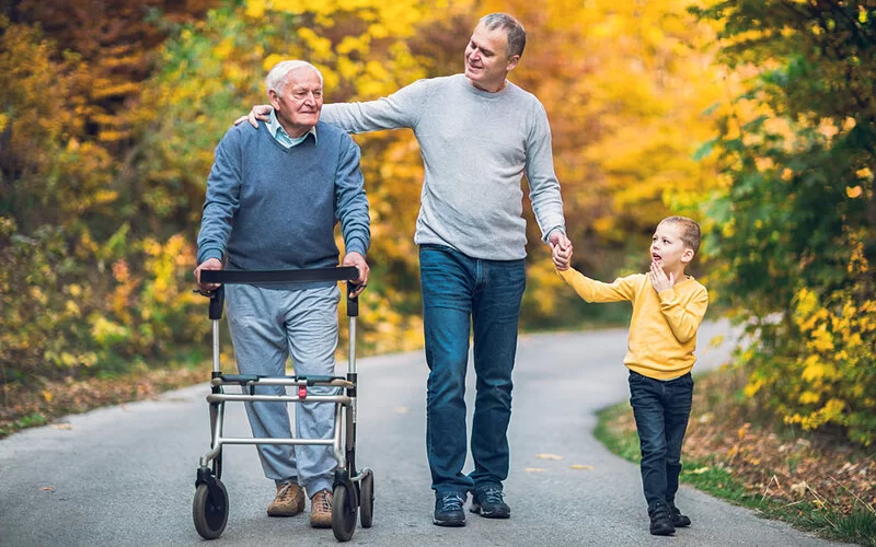 Ein alter Mann mit Rollator geht mit seinem Sohn und seiner Enkelin auf einem gepflasterten Weg durch einen herbstlichen Wald spazieren.