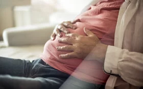 Ein Mann sitzt auf der Couch und hält mit beiden Händen seinen aufgeblähten Bauch.