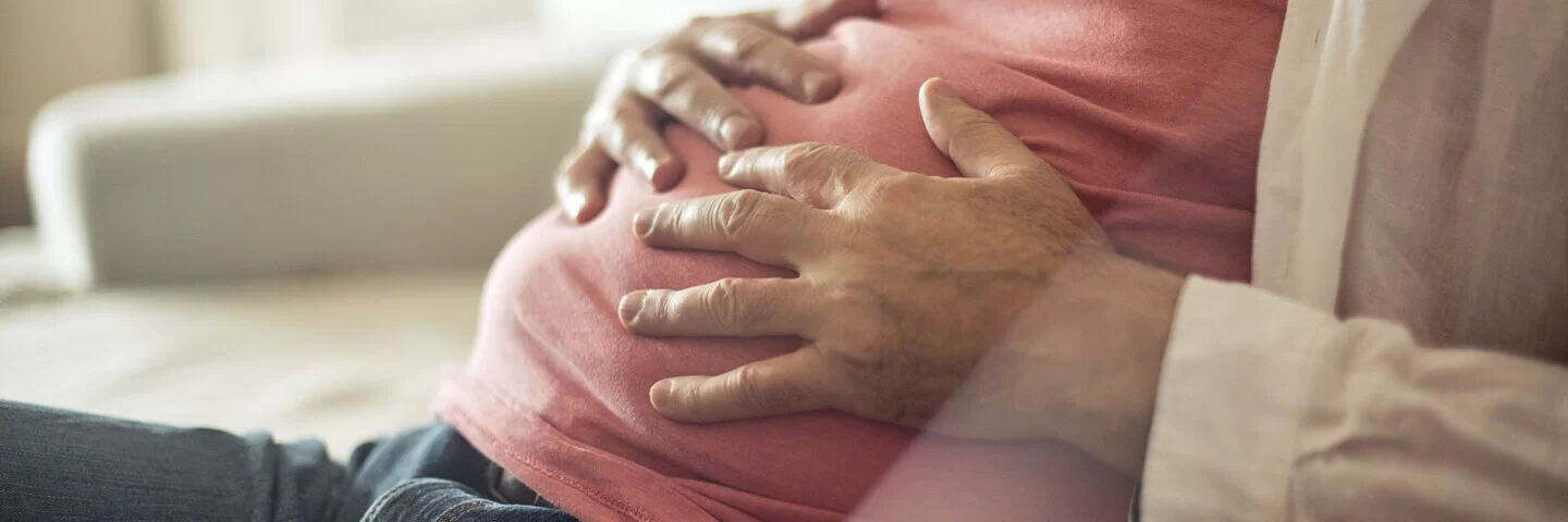 Ein Mann sitzt auf der Couch und hält mit beiden Händen seinen aufgeblähten Bauch.