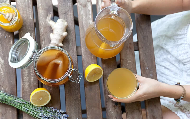 Eine Frau hat sich aus einer Kanne Kombucha in ein Glas gefüllt. Neben der Kanne steht ein geöffnetes Kombucha-Gärgefäß mit SCOBY.