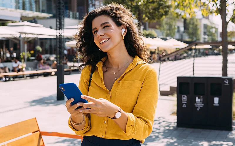 Eine Frau in einer gelben Bluse mit In-Ear-Kopfhörern steht auf der Straße, hält ein Mobiltelefon in ihren Händen und lächelt.