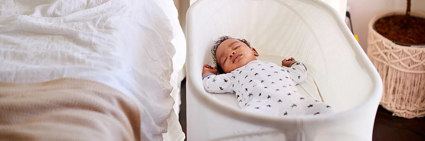 Ein Baby schläft auf dem Rücken in seinem eigenen Bettchen neben dem Elternbett im Schlafzimmer.