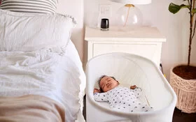 Ein Baby schläft auf dem Rücken in seinem eigenen Bettchen neben dem Elternbett im Schlafzimmer.