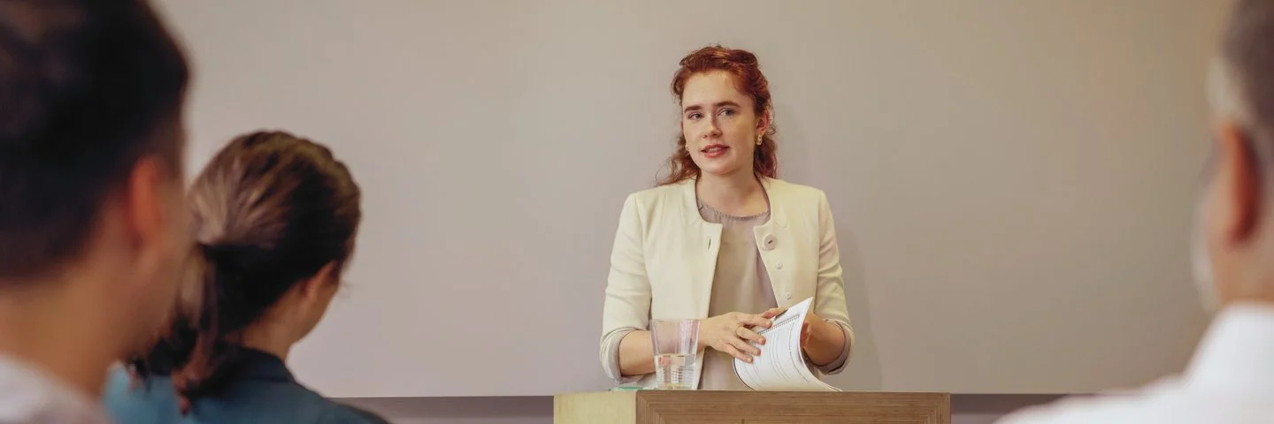 Eine Person steht an einem Rednerpult mit Unterlagen in der Hand und spricht vor mehreren sitzenden Zuhörenden in einem Seminar- oder Vortragsraum.