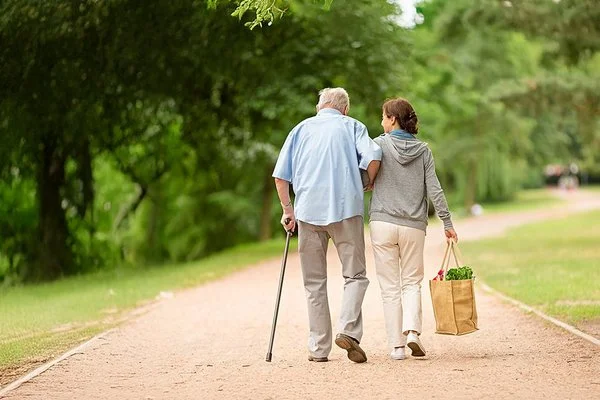 Ein älterer Mann mit Gehstock hält sich am Arm einer Frau fest, die eine Einkaufstasche mit Lebensmitteln trägt. Sie laufen zusammen durch einen Park.