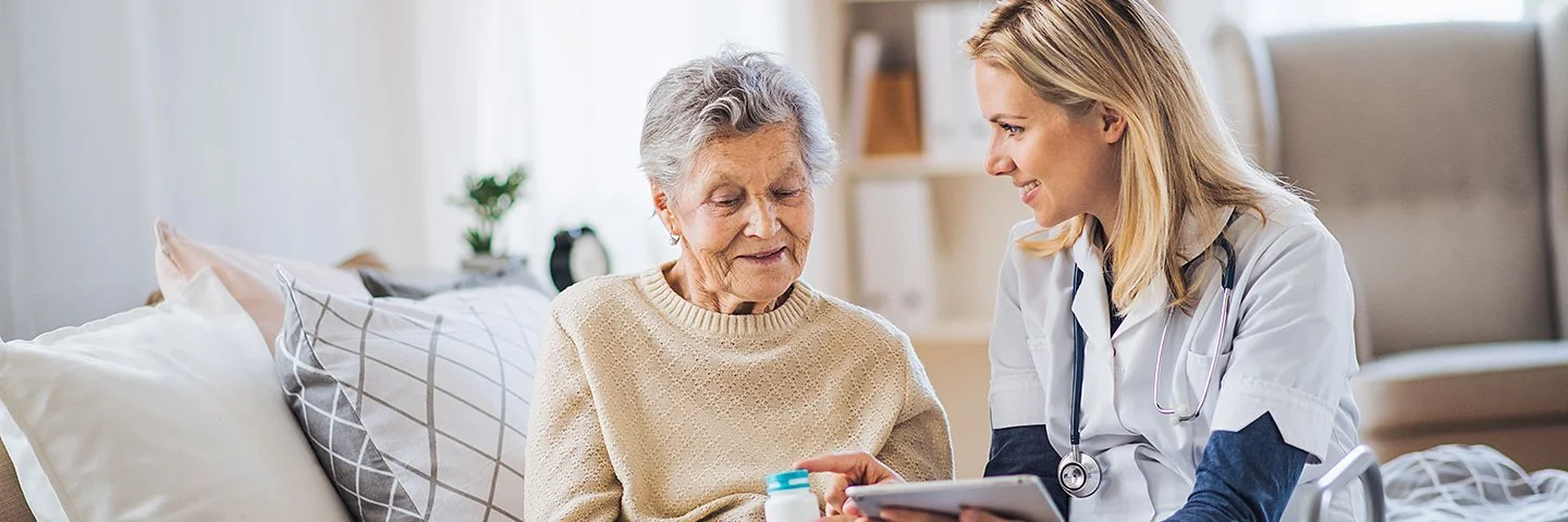 Eine ältere Frau sitzt auf einem Sofa und wird von einer Pflegekraft bei der Einnahme von Medikamenten unterstützt.