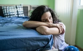 Eine Frau mit Burnout oder Depressionen liegt halb auf dem Bett und schaut mit leerem Blick auf den Boden.