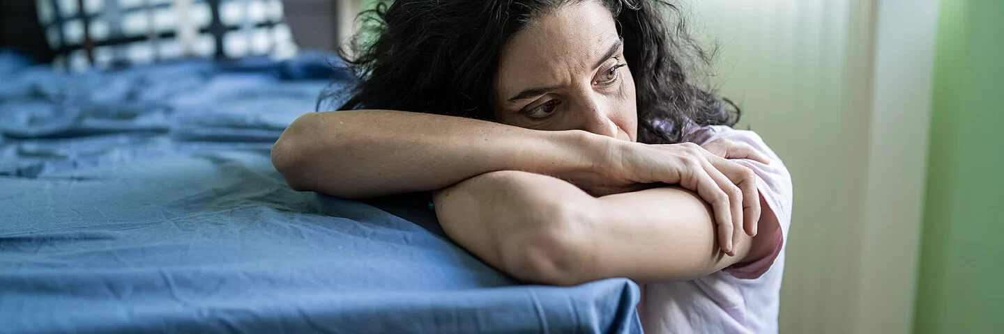 Eine Frau mit Burnout oder Depressionen liegt halb auf dem Bett und schaut mit leerem Blick auf den Boden.