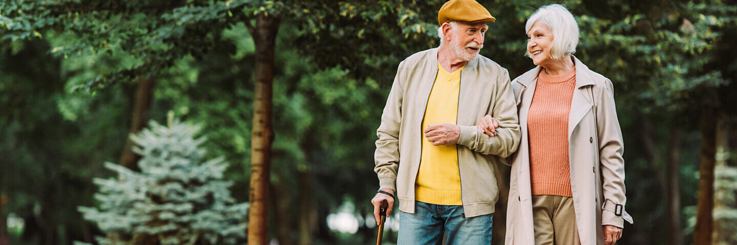 Ein Senior geht gemeinsam mit einer Seniorin durch einen Park. Der Senior hält in seiner linken Hand einen Gehstock und die Seniorin hält sich am rechten Arm des Senioren fest. Sie unterhalten sich und lächeln dabei.