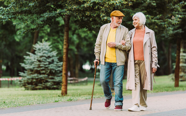 Ein Senior geht gemeinsam mit einer Seniorin durch einen Park. Der Senior hält in seiner linken Hand einen Gehstock und die Seniorin hält sich am rechten Arm des Senioren fest. Sie unterhalten sich und lächeln dabei.