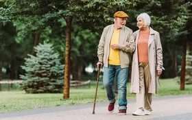 Ein Senior geht gemeinsam mit einer Seniorin durch einen Park. Der Senior hält in seiner linken Hand einen Gehstock und die Seniorin hält sich am rechten Arm des Senioren fest. Sie unterhalten sich und lächeln dabei.