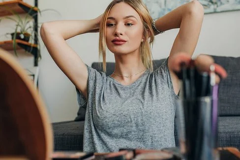 Eine junge Frau sitzt an einem Schminktisch und betrachtet sich im Spiegel. Dabei hält sie sich die Haare aus dem Gesicht.