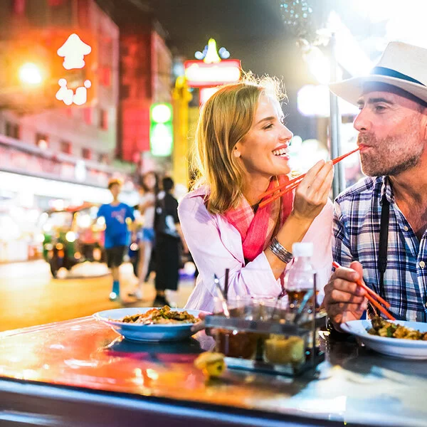 Eine Frau und ein Mann, um die 40 Jahre alt, sitzen nebeneinander in einem chinesischen Street-Food-Restaurant und essen mit Stäbchen. Die Frau gibt dem Mann etwas zum Probieren in den Mund und lacht dabei.