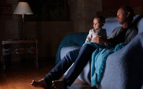 Ein Mann mit postpartaler Depression sitzt mit seinem Baby auf der Couch und schaut Fernsehen.