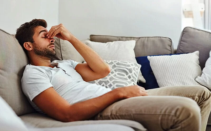 Ein Mann mit Nasennebenhöhlenentzündung hält sich die Nase und sitzt auf der Couch.