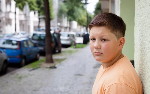 Ein übergewichtiger Teenager lehnt mit dem Rücken an einer Hauswand. Im Hintergrund befinden sich eine städtische Straße, Altbauten, geparkte Autos und Straßenbäume.