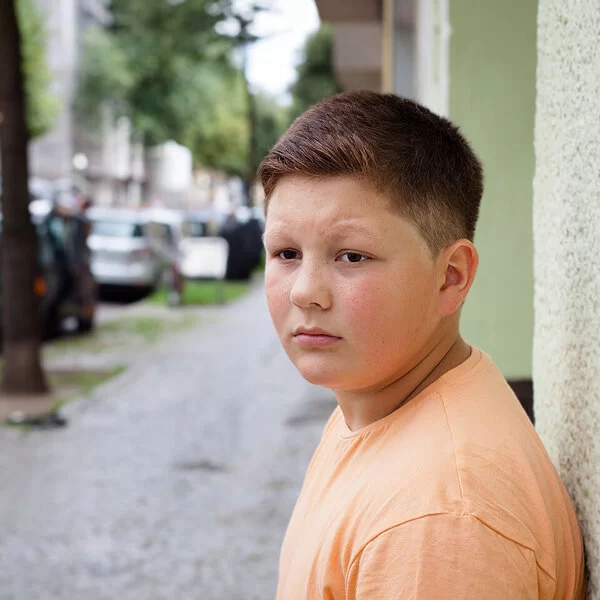 Ein übergewichtiger Teenager lehnt mit dem Rücken an einer Hauswand. Im Hintergrund befinden sich eine städtische Straße, Altbauten, geparkte Autos und Straßenbäume.
