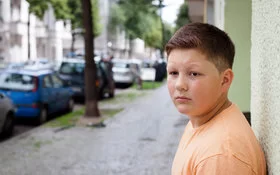 Ein übergewichtiger Teenager lehnt mit dem Rücken an einer Hauswand. Im Hintergrund befinden sich eine städtische Straße, Altbauten, geparkte Autos und Straßenbäume.