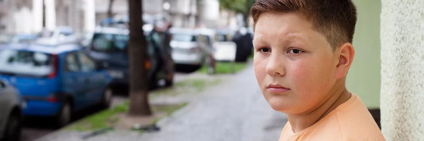Ein übergewichtiger Teenager lehnt mit dem Rücken an einer Hauswand. Im Hintergrund befinden sich eine städtische Straße, Altbauten, geparkte Autos und Straßenbäume.
