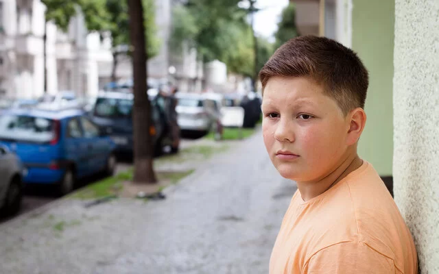 Ein übergewichtiger Teenager lehnt mit dem Rücken an einer Hauswand. Im Hintergrund befinden sich eine städtische Straße, Altbauten, geparkte Autos und Straßenbäume.