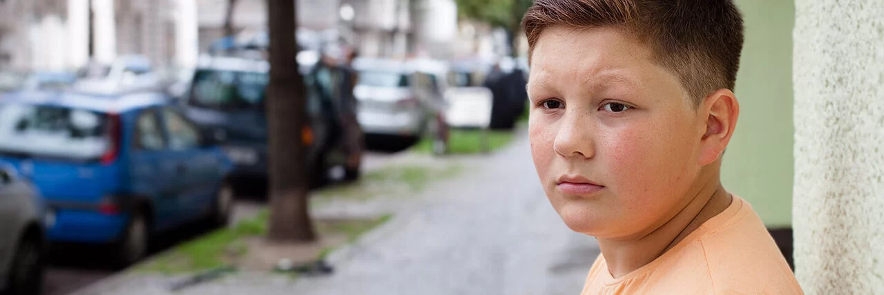Ein übergewichtiger Teenager lehnt mit dem Rücken an einer Hauswand. Im Hintergrund befinden sich eine städtische Straße, Altbauten, geparkte Autos und Straßenbäume.