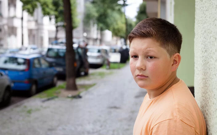 Ein übergewichtiger Teenager lehnt mit dem Rücken an einer Hauswand. Im Hintergrund befinden sich eine städtische Straße, Altbauten, geparkte Autos und Straßenbäume.