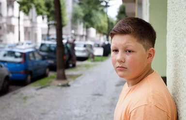 Ein übergewichtiger Teenager lehnt mit dem Rücken an einer Hauswand. Im Hintergrund befinden sich eine städtische Straße, Altbauten, geparkte Autos und Straßenbäume.