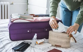 Eine Frau packt Tabletten in eine Kosmetiktasche. Daneben liegt ein geöffneter Koffer auf dem Bett.