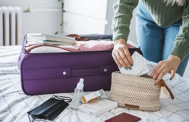 Eine Frau packt Tabletten in eine Kosmetiktasche. Daneben liegt ein geöffneter Koffer auf dem Bett.