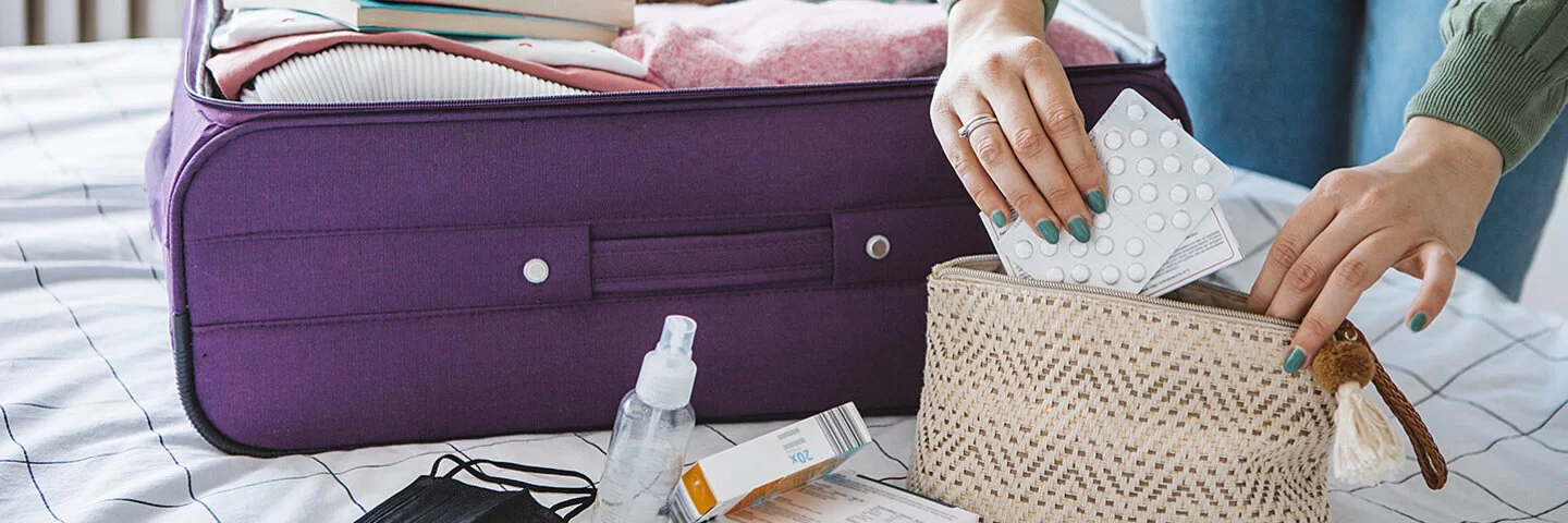 Eine Frau packt Tabletten in eine Kosmetiktasche. Daneben liegt ein geöffneter Koffer auf dem Bett.