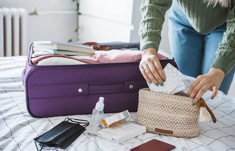 Eine Frau packt Tabletten in eine Kosmetiktasche. Daneben liegt ein geöffneter Koffer auf dem Bett.