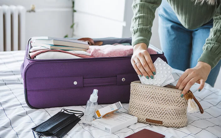 Eine Frau packt Tabletten in eine Kosmetiktasche. Daneben liegt ein geöffneter Koffer auf dem Bett.