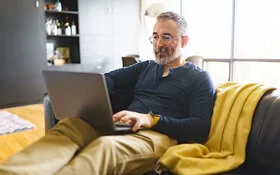 Ein Mann Mitte 50 sitzt entspannt mit einem Laptop auf dem Schoß auf einem Sofa.