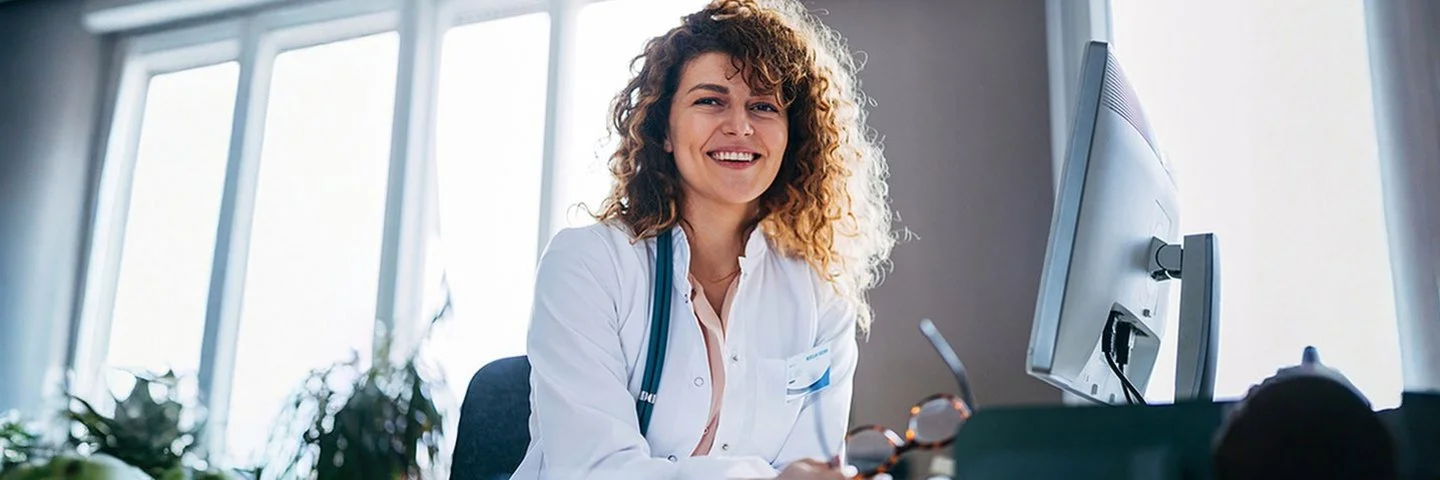 Eine lächelne Ärztin sitzt an ihrem PC und schaut in die Kamera.