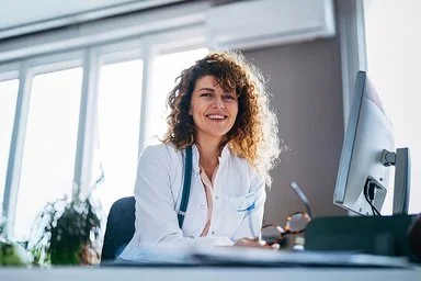 Eine lächelne Ärztin sitzt an ihrem PC und schaut in die Kamera.