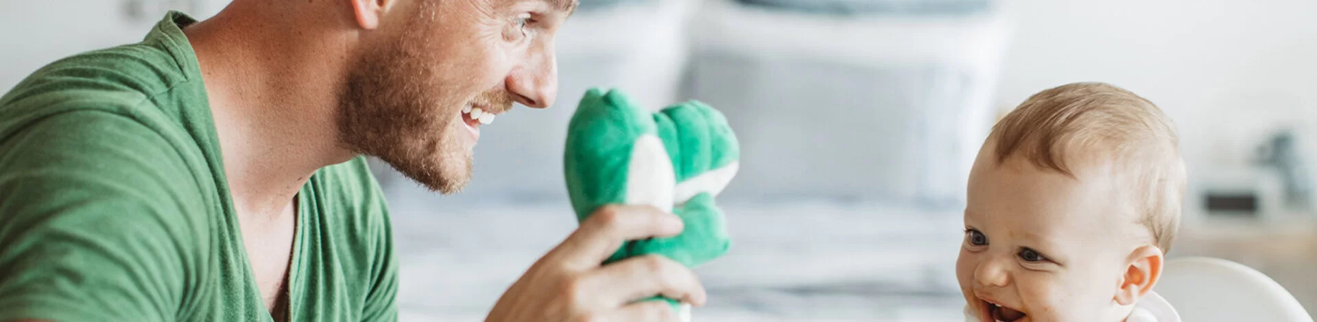Ein Mann lächelt einem Kind im Küchenstühl zu und hält eine Plüschfigur in der rechten Hand.